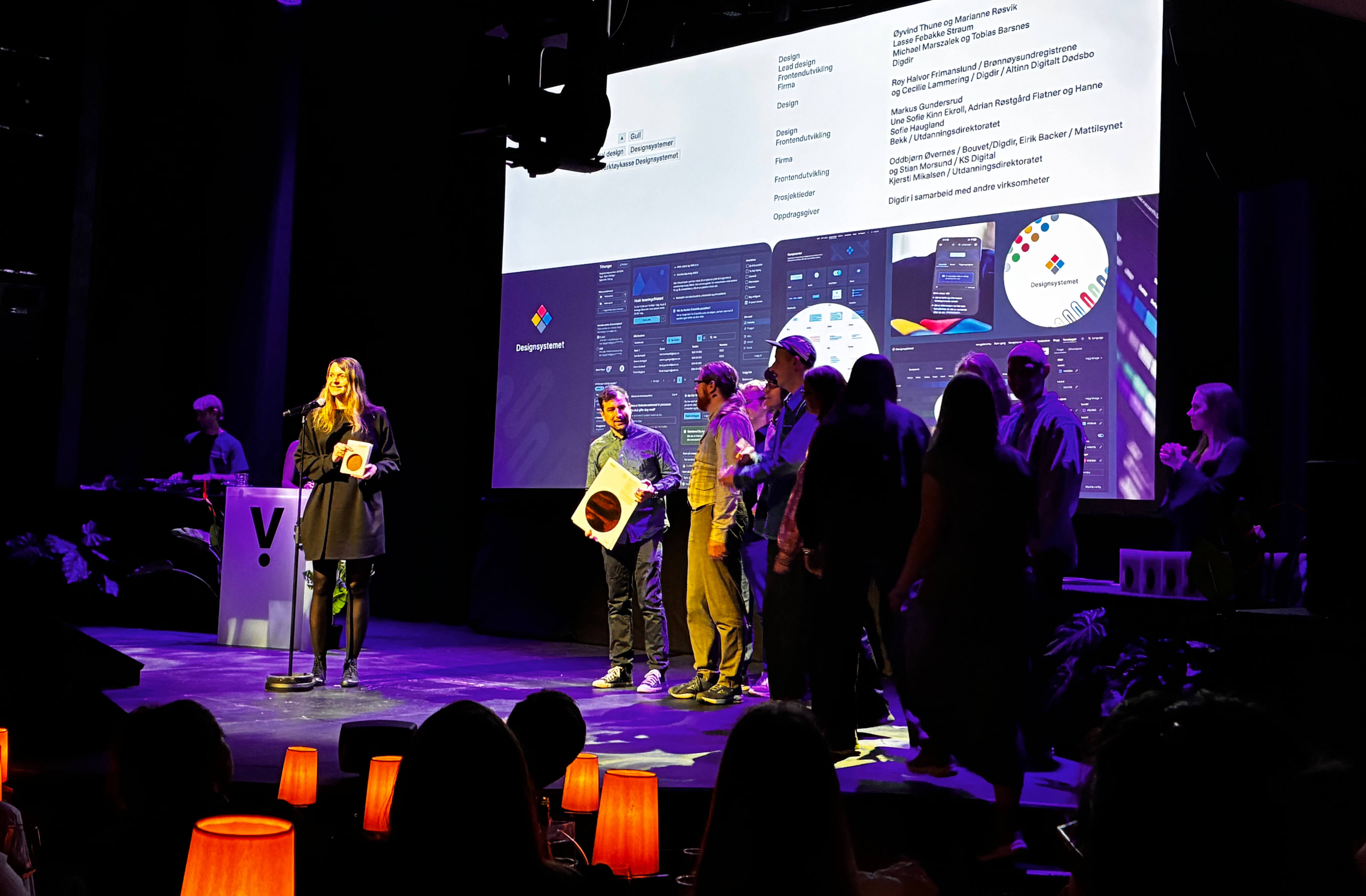 People on stage during the Visuelt award ceremony, with Designsystemet shown on a large screen in the background. Two people are holding awards in front of an audience.
